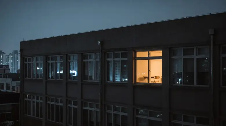 밤의 학교 건물. 캄캄한 창들 사이에 교실 하나만 따뜻하게 불이 켜져 있다.